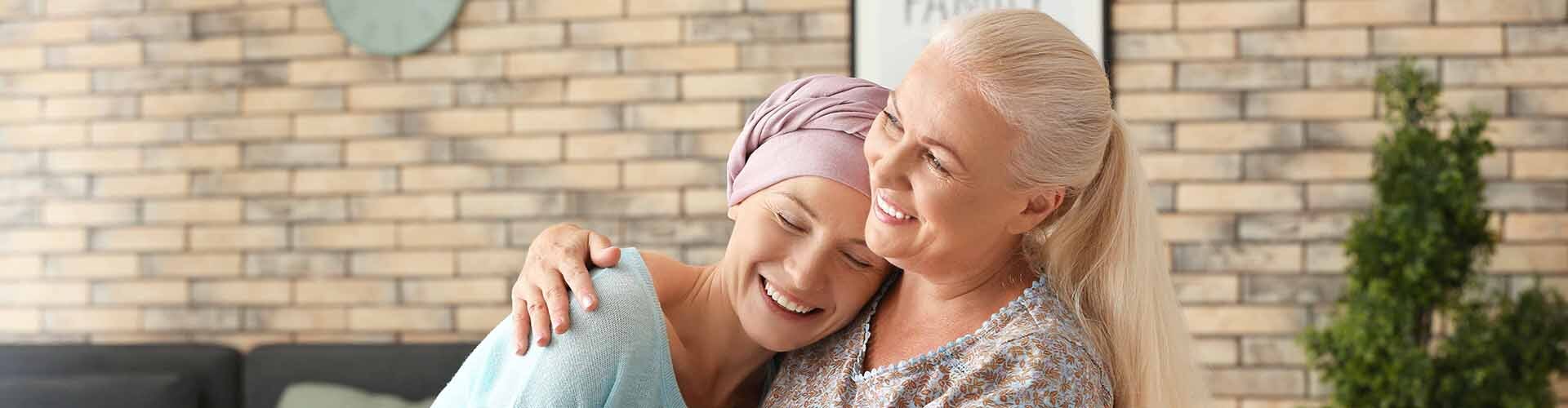 two women sitting on a couch hugging with one wearing a chemo turban
